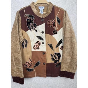 May include: A long-sleeve jacket featuring a color-blocked design in shades of brown, tan, and cream. The front displays embroidered black floral motifs with thin white outlines. It has a round neckline, a button-down front, and dark brown cuffs.