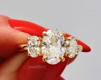 May include: A gold ring featuring three oval-cut diamonds. The central diamond is larger than the two flanking stones. The ring is held against a light gray background. The text "jewellNYC" is visible.