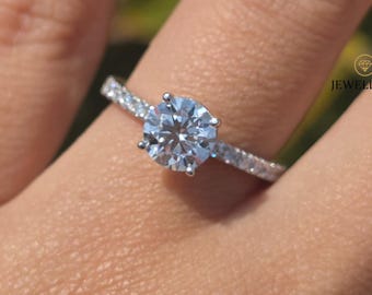 May include: A silver engagement ring with a large, round, clear gemstone as the focal point. The band is encrusted with smaller, clear gemstones. The ring is on a person's finger, with a blurred green background.