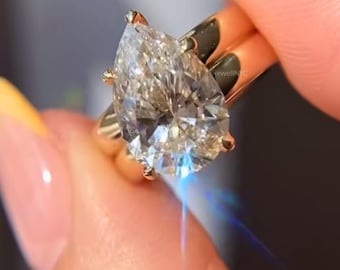 Könnte beinhalten: Nahaufnahme eines goldenen Rings mit einem großen, birnenförmigen Diamanten. Der Diamant ist stark facettiert und reflektiert das Licht. Der Ring hat ein goldenes Band, und der Diamant wird von vier Krappen gehalten.