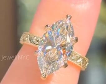 Puede incluir: Un anillo de diamantes con corte marquesa y una banda dorada. El anillo presenta un diseño ornamentado en la banda y un diamante grande y transparente. El anillo está sostenido por un dedo. El texto "jewelNYC" es visible.