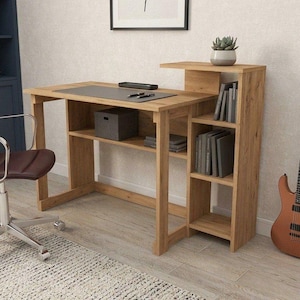 Puede incluir: Escritorio de madera clara con estanterías integradas y una estantería superior elevada. El escritorio tiene una almohadilla gris. Una silla de oficina de cuero marrón y una guitarra son visibles en el fondo.