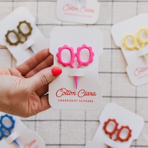 May include: A hand holds a white card with pink embroidery scissors. The scissors have a scalloped edge and a pink handle. The card has the text "Cotton Clara Embroidery Scissors" in red. Other cards with scissors in various colors are in the background.