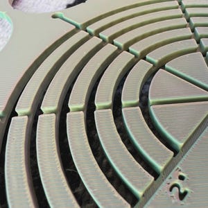 May include: Close-up of a pale green, circular object with a series of curved, segmented ridges. The object has a textured surface and appears to be made of a durable material. The design includes a partial numerical marking.