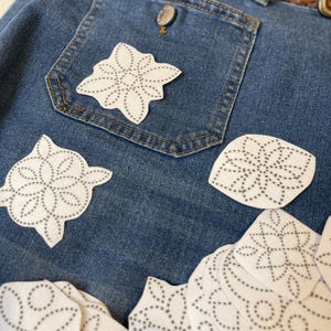May include: Close-up of blue denim jeans with a brown leather belt. White, flower-shaped appliques with gray dotted outlines are scattered on the jeans. The jeans pocket has a single applique attached.