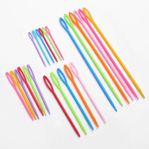 May include: A collection of colorful plastic yarn needles in various sizes. The needles are in shades of pink, orange, yellow, green, blue, and red. The eye of each needle is oval-shaped. The needles are arranged on a white surface.