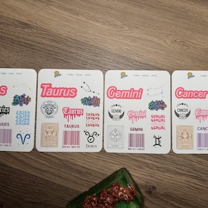 May include: Four white square coasters with colorful zodiac designs. Each coaster features a different zodiac sign: Aries, Taurus, Gemini, and Cancer. The designs include the sign's name, symbols, and dates.