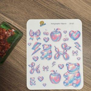 May include: A sheet of holographic stickers featuring various designs, including apples, hearts, bows, lightning bolts, and gummy bears. The stickers have a pastel color scheme of pink, blue, and purple. The text "Holographic Objects LB-23" is printed on the sheet.