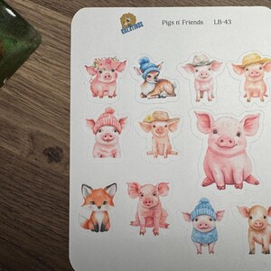 May include: A sheet of stickers featuring watercolor illustrations of pigs and a fox. The pigs are depicted in various poses and outfits, including hats and sweaters. The text "Pigs n' Friends" and "LB-43" are visible.