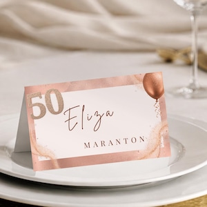 Op de afbeelding: Een roségouden naamkaartje voor een 50e verjaardag. De kaart toont de naam "Eliza" en "Maranton" in elegant schrift, met een ballongrafiek. De kaart ligt op een wit bord met een gouden onderzetter.