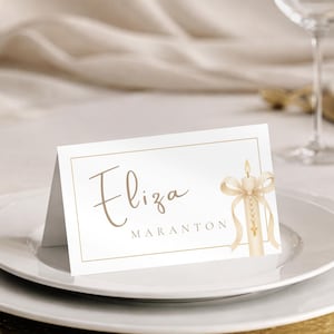 Op de afbeelding: Een witte naamkaart met gouden letters en een kaarsillustratie, geplaatst op een wit bord met een gouden oplader. De kaart leest "Eliza Maranton". Wijnglazen en bestek zijn op de achtergrond te zien.