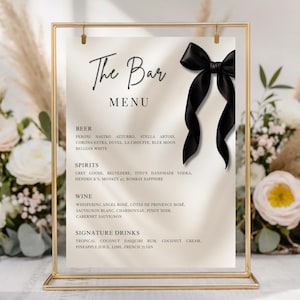 Op de afbeelding: Een goudkleurig omlijst menu voor de bar met een zwart lint. Het menu bevat bier, sterke drank, wijn en speciale drankjes. De tekst is zwart op een crèmekleurige achtergrond. Bloemschikkingen zijn op de achtergrond.