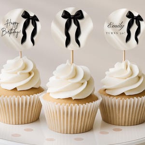 Op de afbeelding: Drie cupcakes met witte frosting en decoraties. De ronde decoraties hebben een zwarte strik en de teksten "Happy Birthday!" en "Emily TURNS 30!". De cupcakes staan op een witte plaat met roze stippen.