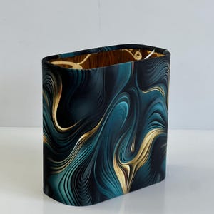 May include: An oval lampshade with a dark teal and gold abstract design. The pattern features flowing lines and swirls, creating a sense of movement. The interior of the lampshade has a warm, wooden appearance.