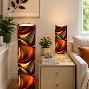 Op de afbeelding: Een hoge vloerlamp en een kleinere bijpassende tafellamp, beide met cilindrische kappen met een levendig, abstract patroon van golvende oranje, rode, blauwgroene en donkerbruine lijnen. Elke lamp staat op drie dunne, witte metalen poten.