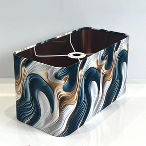 May include: A rectangular lampshade featuring a flowing design in navy, gold, and white. The interior is brown, and the frame is white. This lampshade is designed to fit over a lamp base, adding a decorative touch.