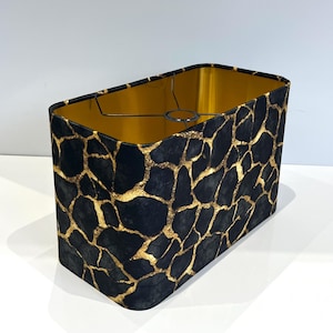 Peut inclure: Abat-jour rectangulaire avec un motif noir et or. L'extérieur présente un motif sombre et texturé avec des accents dorés, tandis que l'intérieur est de couleur or vif. L'abat-jour a une armature métallique et est conçu pour s'adapter à une ampoule.