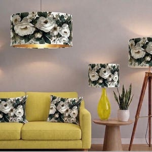 May include: A collection of home decor items featuring a white rose and dark green foliage pattern, including a drum pendant light, a wall sconce, a tripod floor lamp, a table lamp with a yellow base, and two rectangular throw pillows.