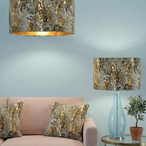 Pode incluir: Um par de candeeiros suspensos e um candeeiro de mesa com abajures a condizer. Os abajures e as almofadas apresentam um padrão abstrato cinzento e dourado. Um sofá rosa e uma pequena mesa lateral com uma planta em vaso completam a cena.