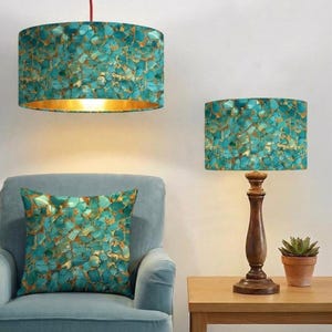 May include: A drum-shaped pendant light and a matching table lamp with a dark wood base, both featuring a turquoise and gold mosaic pattern. A square throw pillow with the same turquoise and gold mosaic design rests on a light blue armchair.