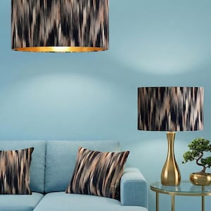 Op de afbeelding: Een moderne woonkameropstelling met een lichtblauwe bank, kussens, een tafellamp en een hanglamp. De lampenkappen en kussens hebben een abstract patroon in zwart, bruin en beige. De lampvoet is goudkleurig.