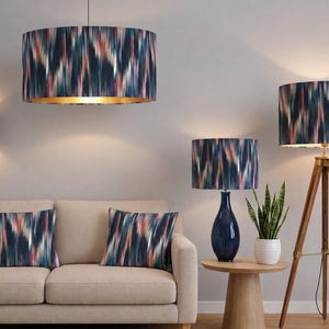 Op de afbeelding: Een collectie verlichting en kussens met een abstract ikat-patroon in donkerblauw, wit en koraal. De set bevat een grote cilindervormige hanglamp, een wandlamp, een tafellamp met een blauwe voet, een driepoot vloerlamp en twee kussens.