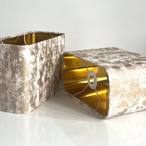 Op de afbeelding: Twee rechthoekige lampenkappen. De buitenkant is een getextureerde, gebroken witte stof met gouden accenten. De binnenkant is goudkleurig. De kappen zijn aan de boven- en onderkant gebogen. De kappen zijn ontworpen om met een lamp te worden gebruikt.