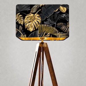 May include: A rectangular lampshade with a black background and gold and black tropical leaf pattern. The lampshade is supported by a wooden tripod base. The lampshade has a gold trim. The lamp is a decorative home decor item.