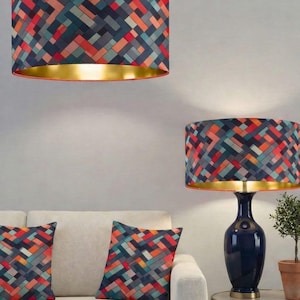 May include: A cylindrical pendant lamp and a matching table lamp with a dark blue ceramic base, both featuring drum shades with a geometric woven pattern in shades of orange, teal, and dark grey, complemented by a metallic gold interior lining.
