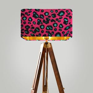 May include: A floor lamp with a rectangular lampshade featuring a pink leopard print with teal and black spots. The lampshade has a gold interior. The lamp is supported by a wooden tripod base with brass accents.