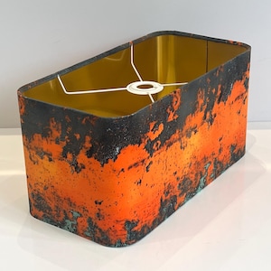 Peut inclure: Abat-jour rectangulaire avec un motif texturé et vieilli. L'extérieur présente un dégradé d'orange et de noir, avec des touches de turquoise. L'intérieur est de couleur or uni, et une armature métallique blanche est visible.