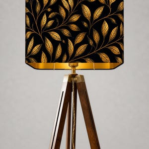 May include: A floor lamp with a rectangular lampshade featuring a black background and gold leaf pattern. The lamp has a wooden tripod base and gold-colored metal accents. The lampshade is approximately 12 inches wide.