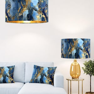 Puede incluir: Pantallas de lámpara y cojines con un diseño abstracto azul y dorado. Las pantallas son cilíndricas, con interior dorado. Los cojines son cuadrados. El patrón presenta colores azules y dorados arremolinados. También se ve una base de lámpara dorada y una mesita auxiliar.