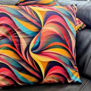 Op de afbeelding: Twee decoratieve kussens met een levendig, abstract ontwerp. De kussens hebben een wervelend patroon van warme kleuren, waaronder rood, oranje, geel en blauw. De kussens liggen op een donkere leren bank.