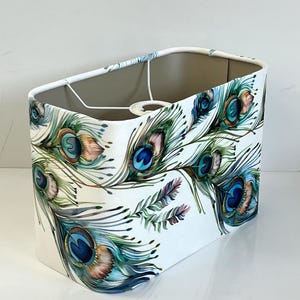 May include: A rectangular lampshade with a white background, adorned with a vibrant peacock feather design. The feathers feature shades of blue, teal, green, and brown. The lampshade has a slightly angled top and a neutral interior.