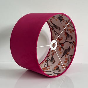 May include: A magenta lampshade with a cylindrical design. The outer surface is a solid magenta, and the inner surface displays a pattern of birds on branches with orange leaves. It has a white metal frame.