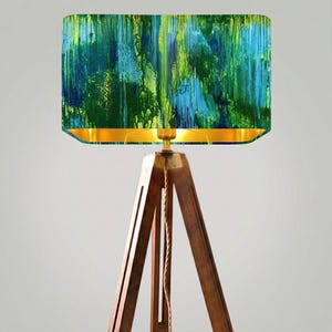 Op de afbeelding: Een moderne vloerlamp met een rechthoekige lampenkap met een abstract ontwerp in groen, blauw en geel. De lampenkap heeft een gouden binnenkant en staat op een donkerbruine houten driepoot. Een decoratief item voor in huis.