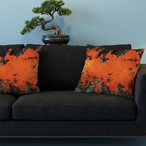 Könnte beinhalten: Zwei quadratische Kissen mit einem abstrakten orange-schwarzen Design liegen auf einem schwarzen Sofa. Ein kleiner Bonsai-Baum in einem dunkelblauen Topf steht auf der Rückseite des Sofas. Der Hintergrund ist eine hellblaue Wand.