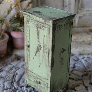 Könnte beinhalten: Ein Miniaturschrank aus Holz, lackiert in blassem Mintgrün mit einem abgenutzten Finish, das darunter liegendes dunkelbraunes Holz freilegt. Das Möbelstück verfügt über eine einzelne hohe Tür mit einem kleinen Knopf und eine untere Schublade.