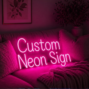 Puede incluir: Un letrero de neón rosa brillante con el texto Custom Neon Sign en una fuente cursiva. El letrero emite un brillo vibrante, iluminando el área circundante con una fuerte luz rosa, creando un punto focal visual distintivo en el espacio.