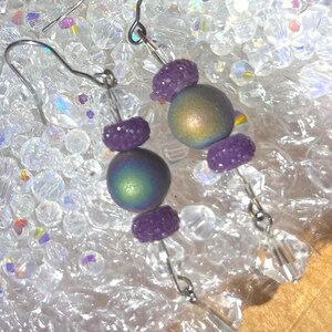 May include: A pair of dangle earrings featuring iridescent, spherical beads in shades of green and gold, accented with small, textured purple beads. The earrings are suspended from silver-tone hooks and adorned with clear, faceted crystal drops.