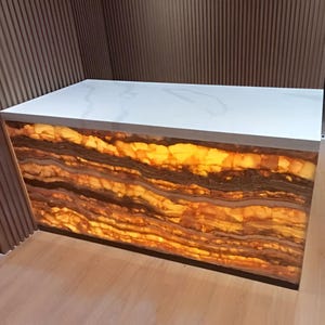 Puede incluir: Una encimera rectangular con una superficie blanca y una base de ónix iluminada y en capas. El ónix muestra bandas de color marrón, naranja y amarillo, creando un efecto cálido y brillante. La encimera está colocada contra una pared de madera.