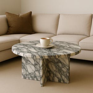 Puede incluir: Una mesa de centro redonda de mármol con un patrón veteado blanco y gris. La mesa tiene una base de pedestal y una taza de té encima. Un cojín marrón descansa sobre un sofá beige en el fondo.