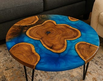 May include: Circular coffee table with a wooden top featuring a blue epoxy resin design. The wood has a natural grain pattern and is surrounded by a vibrant blue hue. The table is supported by black metal hairpin legs.