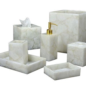 May include: A set of six bathroom accessories in a light, marbled stone. The set includes a tissue box, soap dispenser, wastebasket, tray, toothbrush holder, and soap dish. The dispenser has a gold-colored pump.