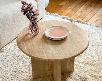 May include: A round, beige travertine coffee table with a cross-shaped base. A small pink glass vase with pink flowers sits on the table, along with two white plates with a pink painted design. The table rests on a cream-colored shag rug.