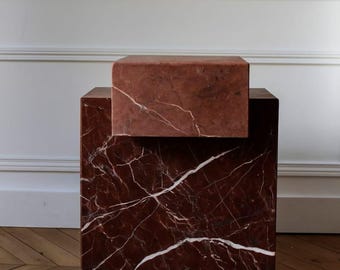 May include: A two-tiered, red marble side table with white veining. The lower section is a cube, and the upper section is a smaller, rectangular block. The table sits on a wooden floor, with a white wall in the background.