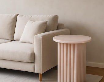 May include: A light pink side table with a round top and vertical, ridged supports. The table is next to a beige sofa with matching pillows. The room has a neutral color palette, with a light rug and a beige wall.