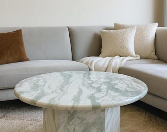 May include: A round coffee table with a white and green marble top and a hexagonal base. The table is placed on a beige rug in front of a gray sofa with decorative pillows. A framed painting hangs on the wall.