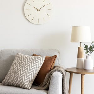 May include: A modern living room setting with a light grey sofa, patterned cushions, and a throw. A wooden side table supports a lamp and vase. A round, pale clock with gold hands is mounted on the wall.